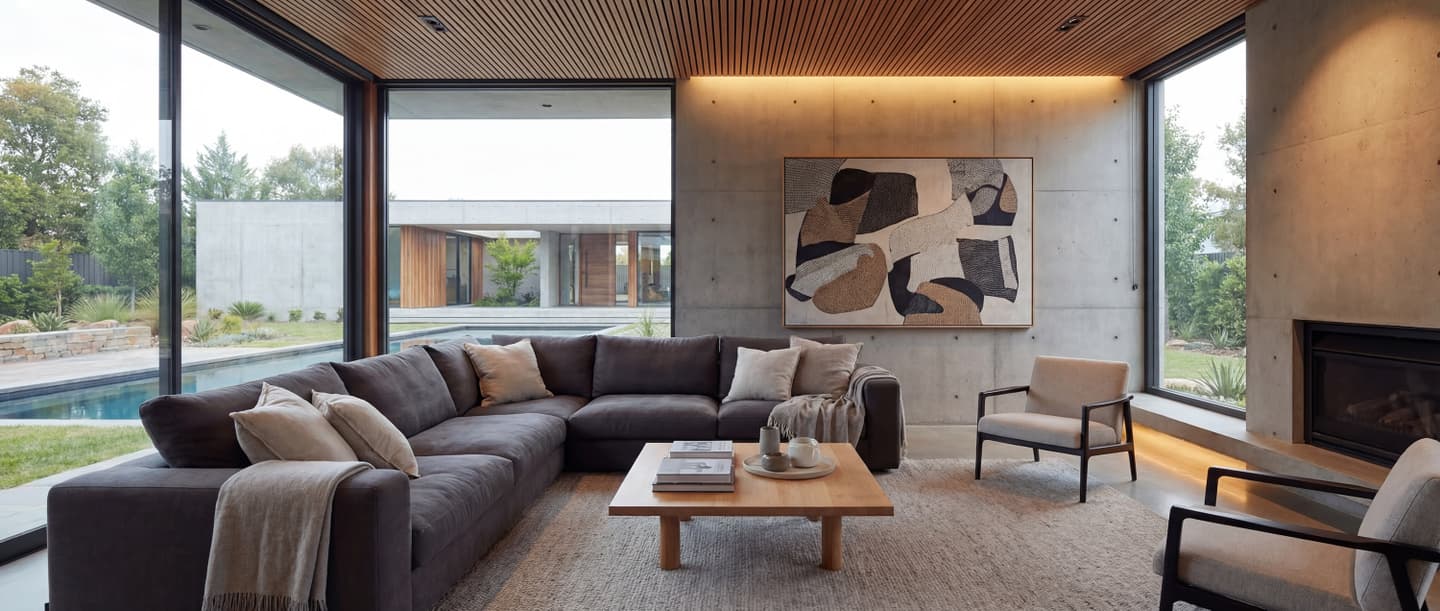 コンクリート打ち放しの壁と木製ルーバー天井が特徴的なモダンなリビングの内観パース。大きな窓からプールのある中庭が見え、グレーのL字型ソファと間接照明が配置された高級感のあるインテリアデザイン。